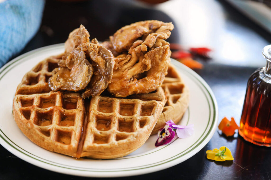 Vegan chicken and waffles made with crispy fried oyster mushrooms on a golden waffle, garnished with edible flowers and served with maple syrup